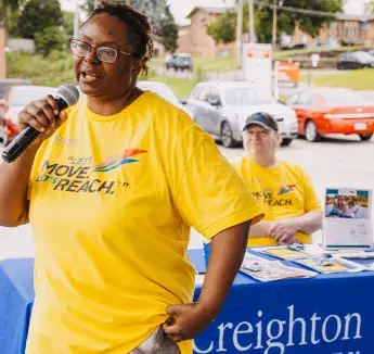 A community health worker in a yellow 'Let's Move Let's Reach' shirt speaks into a microphone at an outdoor health fair.