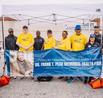 REACH staff and partners hold a banner for the Dr. Frank T. Peak Memorial Health Fair under a white tent.