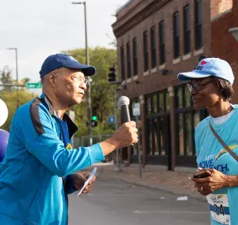 A community member interviews a participant with a microphone during a health event, with blue and purple balloons in the background.