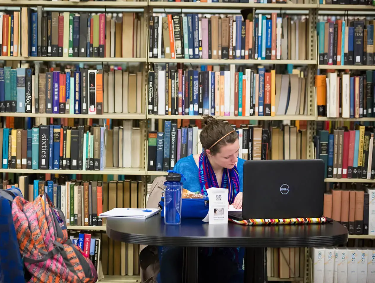 A student studying in the library with book stacks behind them