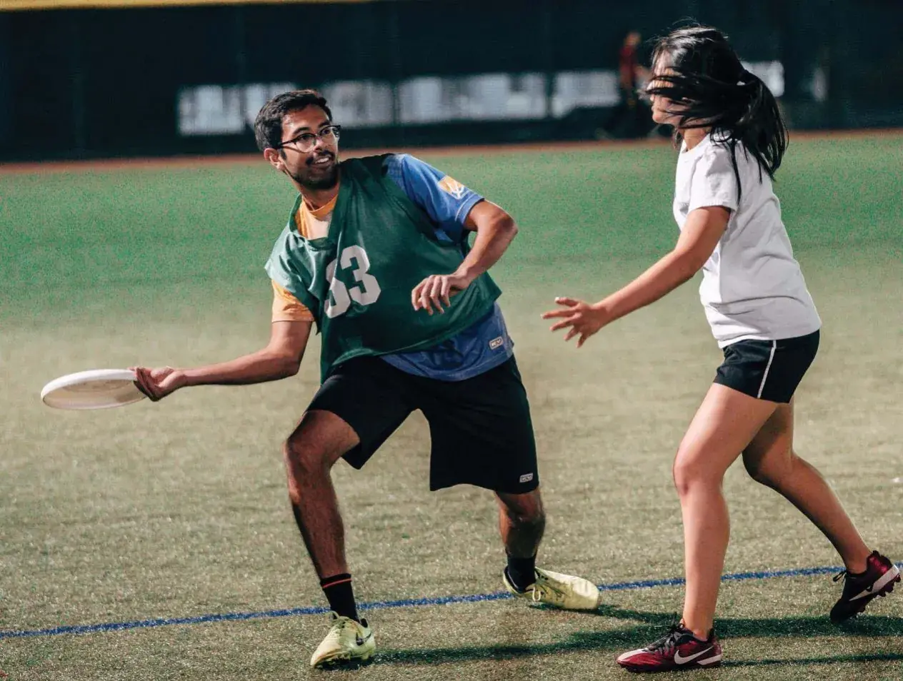 Two students playing frisbee outdoors at night on a lighted field.