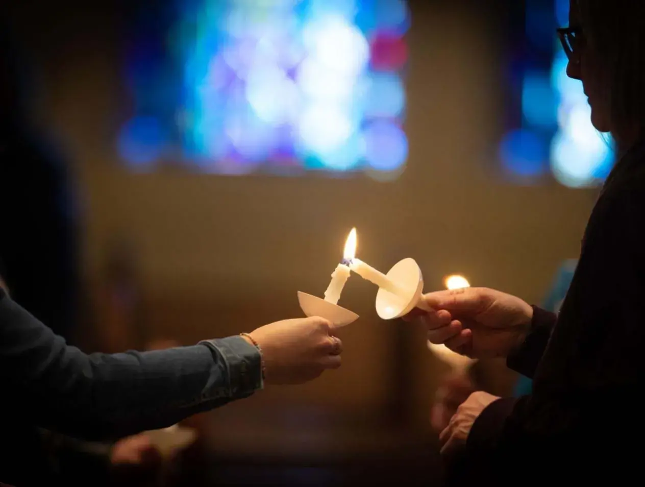 In a dimly lit room, one person uses their taper candle to light another person's candle.