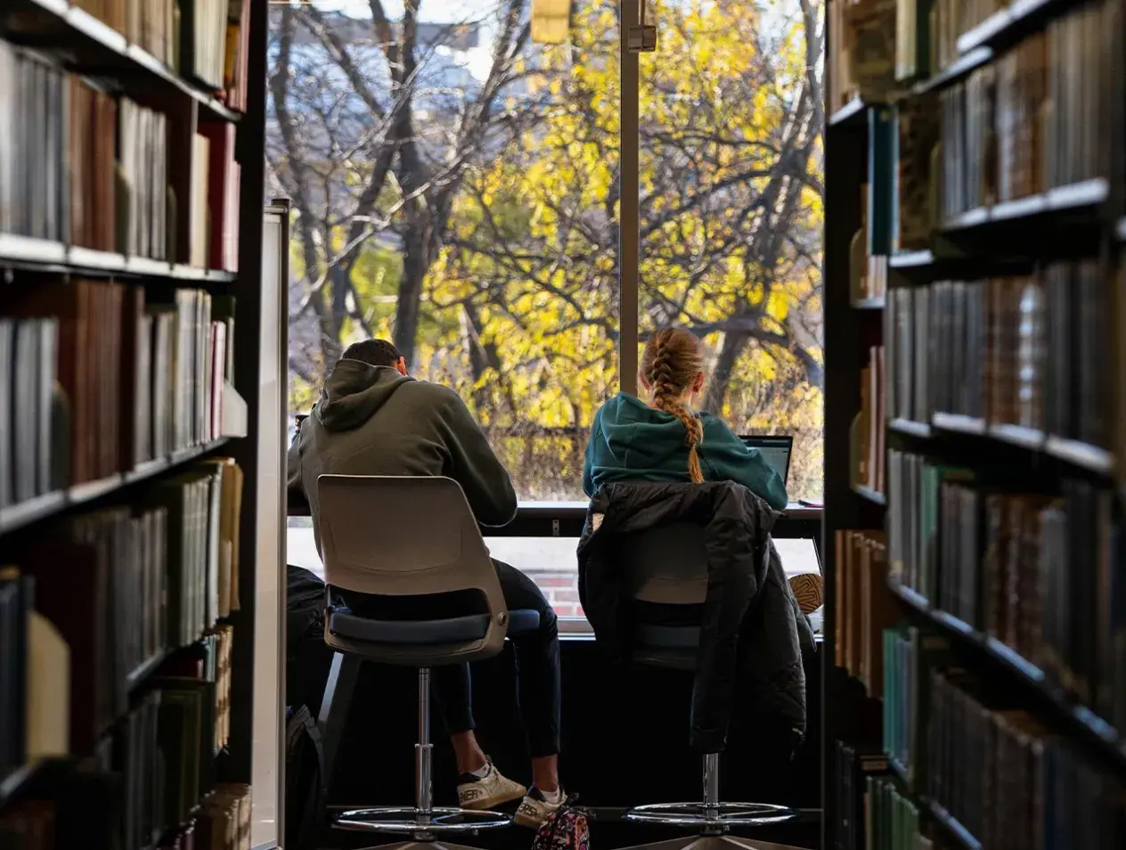 two students sitting in front of window at end of bookshelves