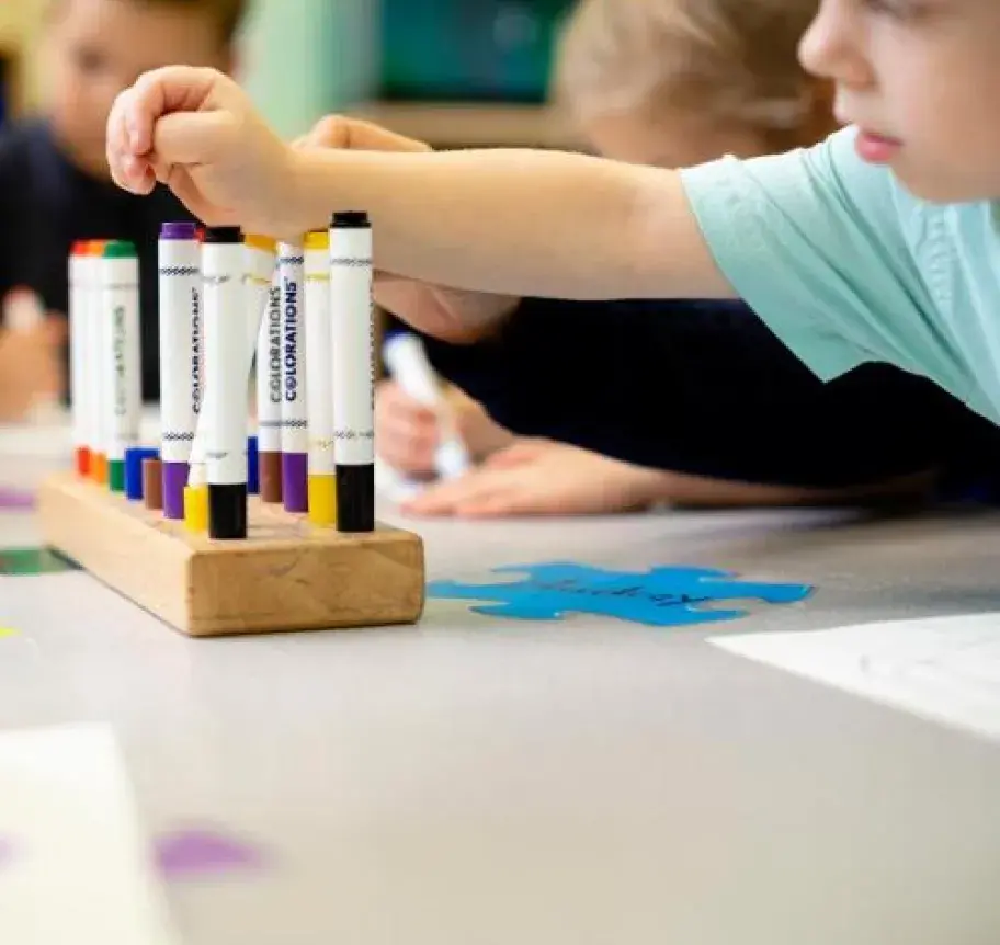 A young child reaches across a table for a marker while coloring